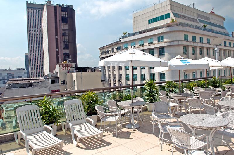 Hotel Atlantico Business Centro Rio de Janeiro Buitenkant foto The photo shows a rooftop area with several white lounge chairs and circular tables. There are umbrellas providing shade over some of the seating. In the background, there are tall buildings, including a modern skyscraper with a distinct architectura